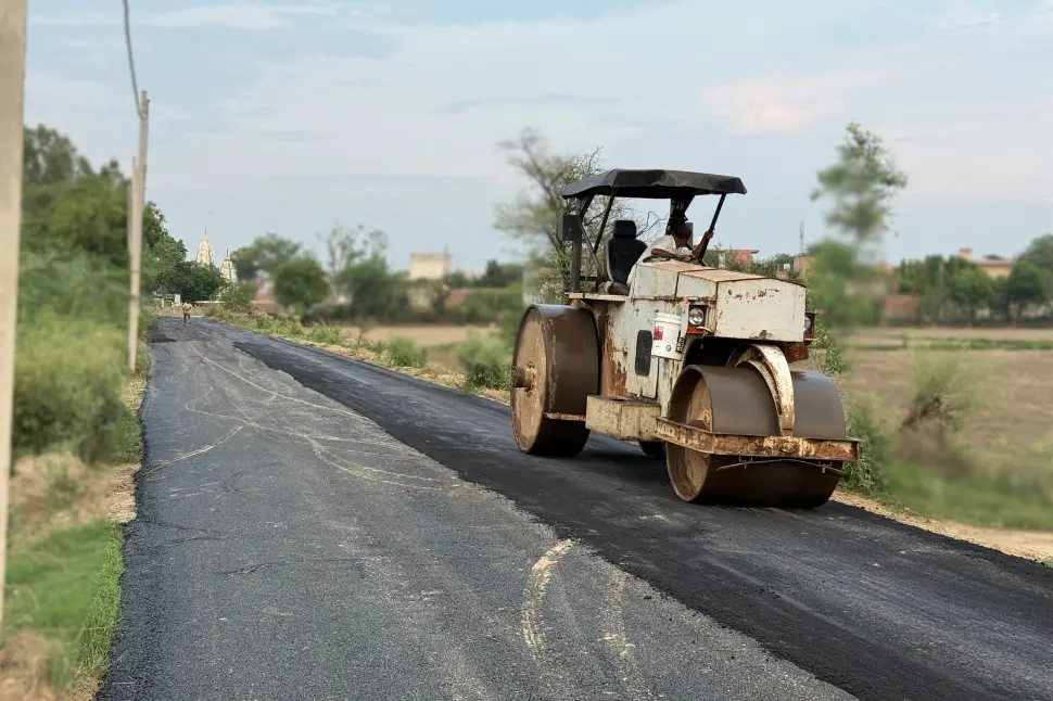 A road roller conducting maintenance on a village road with rural houses and greenery in the background