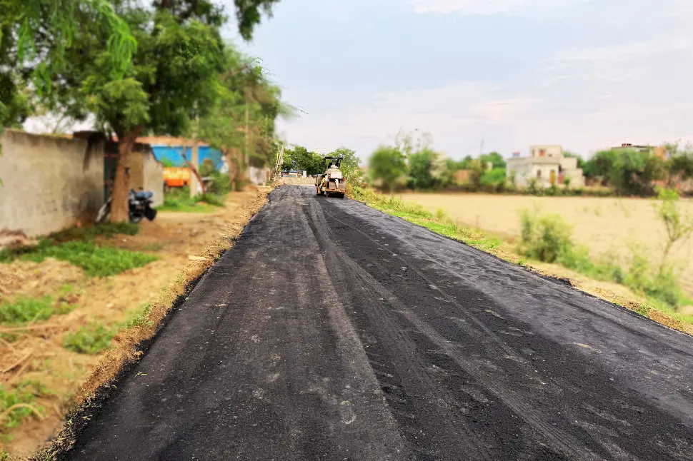 Fresh asphalt road being laid in a rural area with construction equipment and houses visible along the sides